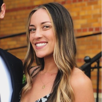 A woman with long, wavy, blonde-highlighted hair smiles at the camera. She is wearing a black and white dress and a gold necklace, standing in front of a brick wall and black railing.