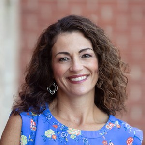 A woman with curly brown hair wearing a blue floral top and dangling earrings smiles at the camera. The background is softly blurred red brick.