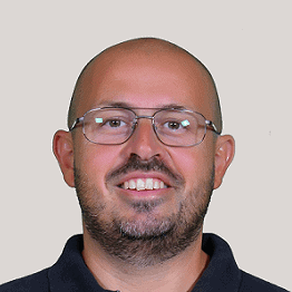 A smiling man with glasses, a beard, and a shaved head, wearing a dark collared shirt, against a plain light background.