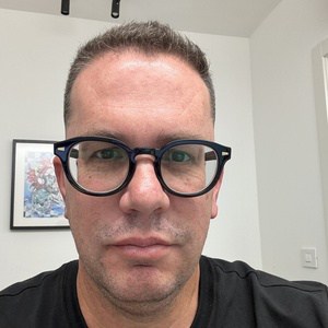 A person with short dark hair and glasses is looking directly at the camera, wearing a black shirt. The background shows a white wall, a framed picture, and a partially visible door.