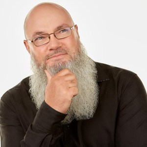 A bald man with glasses and a long gray beard, wearing a black shirt, rests his hand thoughtfully on his chin while smiling slightly against a plain white background.