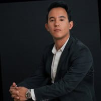 A man with short dark hair wearing a dark suit and white shirt sits with his hands clasped, looking confidently at the camera against a plain dark background.