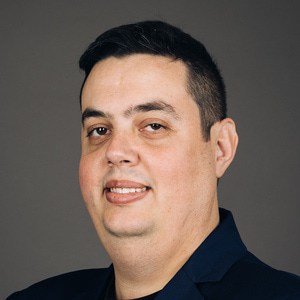 A man with short dark hair, light skin, and brown eyes is smiling slightly. He is wearing a dark blazer and a black shirt, posed in front of a plain dark gray background.