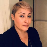 A person with short, light-blonde hair and dark eyebrows, wearing a dark jacket, poses for a photo indoors with a neutral expression. The background features light-colored walls and a door.