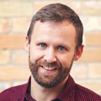 A man with a beard and short hair, wearing a red checkered shirt, is smiling in front of a brick wall.