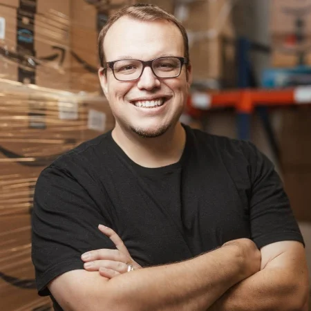 A person with glasses, wearing a black t-shirt, smiles with arms crossed. They stand in front of stacked boxes, suggesting a warehouse or storage setting. The background is slightly blurred.