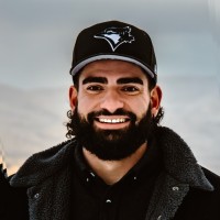 A man with a full beard and mustache, wearing a black Blue Jays baseball cap and a dark jacket, smiles at the camera against a blurred outdoor background.
