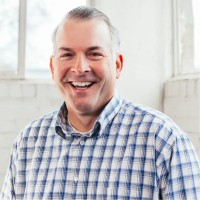 A man with short gray hair and a checked shirt smiles at the camera, standing in front of a white brick wall and large windows with natural light streaming in.