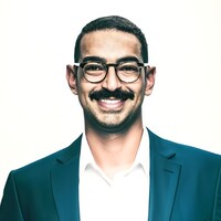 A man with short dark hair, a mustache, and glasses smiles at the camera. He is wearing a dark blue suit jacket over a white dress shirt against a plain white background.