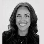 A woman with long, wavy dark hair smiles at the camera. She is wearing a dark top and a small pendant necklace. The background is plain and light-colored.
