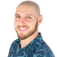 A smiling man with a trimmed beard and a shaved head wears a blue patterned shirt and poses against a white background.