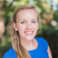 A woman with long blonde hair, wearing a bright blue top and pearl earrings, smiles at the camera with a blurred green outdoor background.