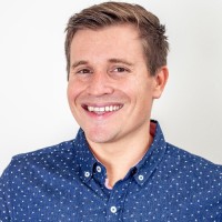 A smiling man with short light brown hair wearing a blue button-up shirt with small white polka dots stands in front of a plain white background.