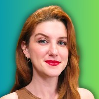 A woman with long red hair and red lipstick, wearing a brown top and small hoop earrings, stands in front of a green and blue gradient background, smiling slightly at the camera.