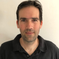 A man with short dark hair and light skin is wearing a black collared shirt and is standing against a plain white background, looking directly at the camera with a neutral expression.