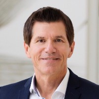 A man with short dark hair is smiling slightly at the camera. He is wearing a dark suit jacket with a white collared shirt. The background is softly blurred and light-colored.