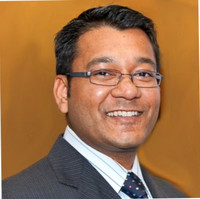 A man with short dark hair wearing glasses, a dark striped suit, a white shirt, and a dotted tie, smiling in front of a plain, warm-toned background.