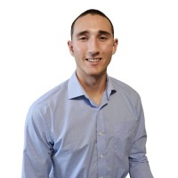 A person with short dark hair wearing a light blue button-up shirt smiles against a plain white background.