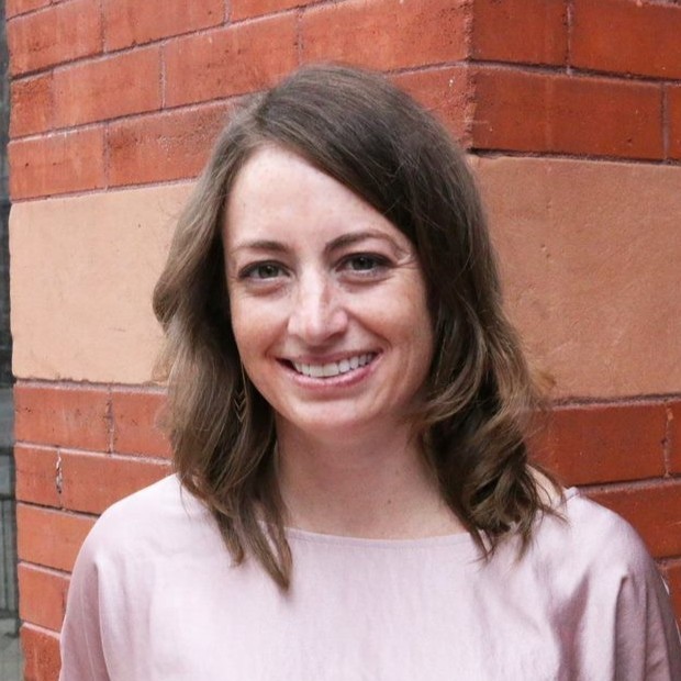 A woman with shoulder-length brown hair smiles in front of a red brick wall. She is wearing a light pink top and is standing outdoors.