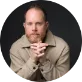 shane barker A man with a trimmed beard and short hair sits leaning forward, hands clasped in front of his face, wearing a beige jacket against a dark background.