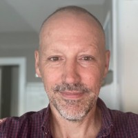 A middle-aged man with a shaved head, trimmed beard, and plaid shirt smiles softly while standing indoors in a well-lit room with a white doorway in the background.