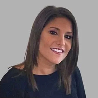 A woman with straight, shoulder-length brown hair and a side part smiles at the camera. She is wearing a black top and is posed against a plain, light gray background.