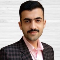 A man with short dark hair and a mustache, wearing a checkered blazer over a light pink collared shirt, poses in front of a light-colored wooden background.