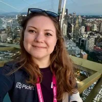 A woman with long brown hair smiles at the camera while taking a selfie on a high outdoor observation deck, with a city skyline and tall buildings visible in the background at dusk.