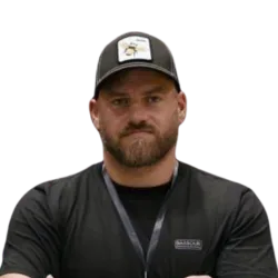 A bearded man wearing a black baseball cap and black t-shirt stands with arms crossed, looking forward with a serious expression.