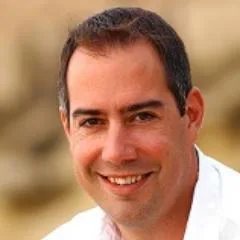 A man with short dark hair and a light complexion is smiling at the camera. He is wearing a white shirt and is photographed against a blurred, neutral background.