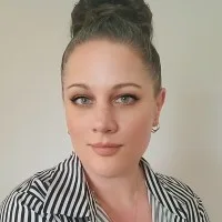 A woman with light eyes and light brown hair styled in a high bun, wearing a striped shirt and small hoop earrings, poses in front of a plain light background.