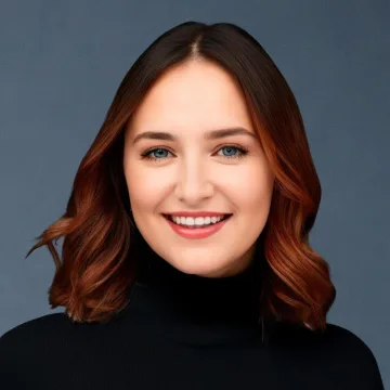 A young woman with shoulder-length brown hair and blue eyes smiles at the camera. She is wearing a black turtleneck sweater and is posed against a plain, dark background.