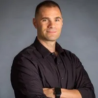 A man with short hair wearing a dark long-sleeve shirt stands with arms crossed, looking confidently at the camera against a plain gray background.