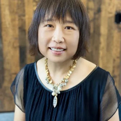 A woman with short dark hair, wearing a sheer black top and a gold statement necklace, smiles at the camera. The background is wooden and softly blurred.