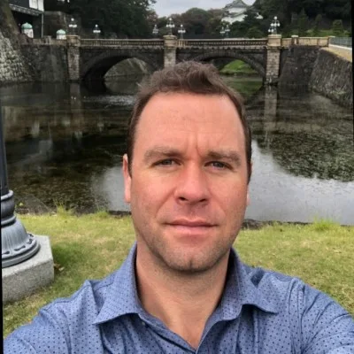 A man in a blue shirt takes a selfie in front of a stone bridge and moat, with trees and a historical building in the background.