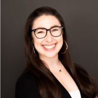 Woman with long dark hair, wearing glasses, hoop earrings, a black blazer, and a white top, smiling in front of a plain dark background.