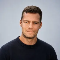 A man with short brown hair, wearing a dark crew-neck sweater, is looking at the camera with a neutral expression. The background is plain and light-colored.
